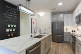 Located on Halcyon Way in Alpharetta, GA, this thoughtfully designed single-floor residence offers a cohesive blend of modern finishes and comfortable living spaces. The open-concept kitchen is a standout feature, showcasing a large waterfall-edge quartz island, soft gray shaker cabinetry, a classic tile backsplash, and a full suite of stainless steel appliances including a French door refrigerator, range, microwave, and dishwasher. Light wood-look flooring runs throughout, enhancing the bright, airy feel. The primary bathroom is elegantly appointed with large-format gray tile flooring, floor-to-ceiling tiled shower walls, a dual-sink vanity with quartz countertops, and an illuminated frameless mirror. The bedrooms offer plush carpeting and generous natural light through wide, double-pane windows dressed with sheer striped curtains. One bedroom features a calming blue-gray accent wall, while another is bathed in soft whites for a serene atmosphere. The living area is anchored by a dramatic dark accent wall adorned with a curated gallery of framed artwork, complemented by pendant Edison bulb lighting and a sectional sofa, creating a warm and inviting gathering space. The overall design palette of whites, grays, and warm wood tones gives this residence a clean, contemporary character throughout.
