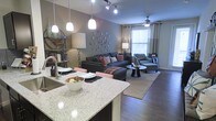 This single-floor residence showcases a thoughtfully designed open-concept layout that seamlessly connects the kitchen and living areas. The kitchen features a generous granite-topped island with seating, served by elegant pendant lights that hang at varying heights, creating a warm and inviting focal point. A undermount sink and modern fixtures complete the functional yet stylish kitchen space.

The living area extends naturally from the kitchen, anchored by dark hardwood flooring that runs throughout. A ceiling fan with integrated lighting presides over the living space, which benefits from natural light streaming through large windows dressed with layered curtains and blinds. The room comfortably accommodates a substantial sectional sofa and an accent chair with ottoman, arranged around a round coffee table atop a patterned area rug.

Warm gray walls provide a neutral backdrop, accented by decorative circular wall art and track lighting overhead. The open flow between the kitchen and living areas creates a sense of spaciousness, making the layout ideal for both everyday living and entertaining. The overall ambiance balances contemporary design with comfortable, livable warmth.