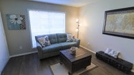 This single-floor space offers a comfortable and well-appointed living environment designed with both function and style in mind. The living area serves as the focal point of the space, showcasing dark hardwood-style flooring that provides a rich contrast against the warm, neutral-toned walls. A large window fitted with horizontal blinds allows generous amounts of natural light to fill the room, creating an open and airy atmosphere. The space is furnished with a plush teal sofa accented by decorative floral pillows, complemented by a distinctive dark wooden trunk-style coffee table mounted on casters, which adds a touch of industrial character to the room. A multi-arm floor lamp positioned in the corner provides additional ambient lighting for evening use. Clean white baseboards frame the room, adding a crisp finishing detail to the overall aesthetic. The layout feels thoughtfully organized, offering a sense of comfort and practicality. The neutral color palette throughout ensures a versatile backdrop suitable for a variety of décor styles. Overall, the space presents a warm, inviting atmosphere that balances everyday livability with tasteful design choices.