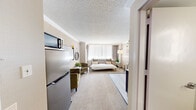 This thoughtfully designed unit offers a seamless single-floor layout that maximizes every square foot. The main living area serves as both a bedroom and lounge space, featuring wall-to-wall carpeting, crown molding, and a textured ceiling that adds depth and character. A large window spanning nearly the full width of the room fills the space with natural light and offers views of the surrounding cityscape, dressed with sheer curtains and draping panels for privacy and ambiance.

The bedroom area is anchored by a full-sized bed with stylish accent pillows, complemented by a cozy seating area with an olive-green armchair and a side table. A wall-mounted flat-screen TV, a stainless steel mini-refrigerator, and a microwave are neatly arranged near the entrance, providing essential in-unit conveniences. A bistro-style high table with wooden stools adds a charming dining nook, while an arc floor lamp provides warm, ambient lighting.

The transition from carpet to hardwood-style flooring near the entrance creates a natural delineation of zones. The bathroom is clean and modern, featuring a glass-enclosed walk-in shower with white tile surround, a round vessel sink on an open vanity with a backlit mirror, and a built-in dark wood shelving unit that adds both storage and visual interest.