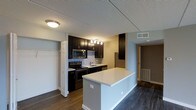 This single-floor residence showcases a well-appointed, open-concept kitchen that serves as the focal point of the space. The kitchen features rich, dark espresso cabinetry extending from floor to ceiling, providing generous storage capacity. A decorative mosaic tile backsplash adds visual texture and contrast against the dark cabinets, while stainless steel appliances — including a built-in microwave, electric range, and refrigerator — offer a sleek, modern aesthetic. A white quartz peninsula island anchors the space, offering additional prep area and casual seating potential. Track lighting illuminates the workspace effectively. Dark hardwood-style flooring runs throughout, creating a cohesive and contemporary look. Adjacent to the kitchen, a closet with a built-in shelving and hanging rod system provides practical storage. A hallway visible in the background suggests additional rooms beyond the main living area. The textured ceiling and neutral wall tones provide a clean backdrop, allowing the bold cabinetry and flooring to stand out as the defining design elements of this functional and modern space.
