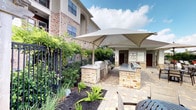 This outdoor kitchen and aquatic amenity area offers a well-appointed al fresco experience designed for both relaxation and entertaining. The outdoor kitchen stations are constructed with handsome stone masonry bases topped with granite countertops, housing built-in stainless steel grills and prep surfaces. Large cantilever shade umbrellas provide generous cover over the cooking and dining areas, creating a comfortable environment even on sunny days. The paver-laid patio flows seamlessly between the kitchen stations and the surrounding landscape, which features lush greenery, ornamental ferns, and manicured shrubs that lend a tropical ambiance to the space. Adjacent to the kitchen area, the expansive resort-style swimming pool takes center stage, showcasing vivid turquoise water, decorative fountain features, and a broad surrounding deck furnished with lounge chairs and seating areas. A grand clubhouse structure with a pitched roof and large windows serves as the architectural backdrop, reinforcing the upscale character of the amenity. Umbrella-shaded lounge groupings and additional seating are positioned poolside, completing the cohesive outdoor living environment under open skies.