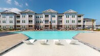 Located at 3868–3898 Cimarron Blvd in Corpus Christi, Texas, this single-floor residence is part of a multi-story community building characterized by a classic coastal architectural style, featuring light siding, multiple balconies, and large windows that allow ample natural light throughout. The community's centerpiece is a striking resort-style pool surrounded by a spacious sundeck furnished with contemporary lounge seating, ideal for the South Texas climate.

Inside, the unit features an open-concept living and kitchen area anchored by warm wood-look flooring that flows seamlessly throughout the main living spaces. The kitchen is equipped with stainless steel appliances, dark shaker-style cabinetry, a subway tile backsplash, pendant lighting over a central island with bar seating, and light stone countertops. The living area is generously proportioned with well-defined wall space and a welcoming, coastal-inspired ambiance.

A hallway connects the living area to the bedroom, which features plush carpeting, a ceiling fan, and a glass-paneled door fitted with blinds for light control. The corridor areas are cleanly finished with neutral tones, crown molding details, and convenient access to additional rooms. Throughout the unit, the design maintains a cohesive, light, and airy aesthetic well-suited to coastal living.