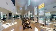 This fitness center is a comprehensive, well-appointed facility designed to support a full range of workout routines. The space is highlighted by a striking cycling studio with soaring double-height ceilings, polished hardwood floors, and expansive floor-to-ceiling windows that flood the room with natural light and offer views of an outdoor pool area framed by palm trees. A row of stationary cycling bikes lines the studio, making it ideal for group or individual spin sessions.

The cardio zone is generously stocked with a row of TRUE-brand elliptical trainers and treadmills, positioned along an open railing that overlooks the cycling area below, creating a sense of connectivity throughout the facility. Arched windows bring in additional natural light, contributing to an open and energizing atmosphere.

The strength training area is equally robust, featuring a wide selection of cable machines and resistance equipment arranged across durable rubber flooring. Full-wall mirrors amplify the sense of space and allow for proper form monitoring during workouts. StairMaster steppers and additional elliptical machines round out the cardio offerings in this zone.

Throughout the facility, recessed lighting complements the natural light, ensuring a bright and welcoming environment. The overall design is clean, modern, and functional, offering users everything needed for a thorough and effective fitness experience.