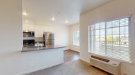 This single-floor unit offers a thoughtfully laid-out living space with a clean, neutral aesthetic that creates a bright and welcoming atmosphere throughout. The open-concept kitchen and living area serves as the heart of the unit, where the kitchen features white cabinetry with chrome hardware, dark countertops, and a full suite of stainless steel appliances including a refrigerator, microwave, and range. The kitchen peninsula provides additional workspace and transitions seamlessly into the living area, which is finished with plush carpeting and benefits from generous natural light streaming through large windows fitted with white blinds. A wall-mounted HVAC unit ensures year-round climate comfort in this shared living space. The bedroom is a well-proportioned, carpeted room with soft, neutral walls, offering a calm and restful environment. A closet with double doors provides convenient storage just off the entry. The bathroom is finished with warm stone-toned tile flooring and includes a full tub and shower combination, toilet, and vanity. Wood-look flooring in the kitchen corridor adds warmth and is easy to maintain. Overall, the unit presents a functional, move-in-ready layout with consistent finishes and ample natural light throughout.