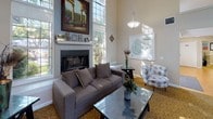 This welcoming single-story clubhouse is designed to serve as a central gathering hub, combining elegant architectural details with comfortable, functional furnishings throughout. The space is immediately defined by its dramatic vaulted ceilings, which rise to impressive heights and are complemented by large transom and floor-to-ceiling windows that flood the interior with natural light and offer views of the surrounding greenery.

The main open area is anchored by a patterned gold-toned carpet that adds warmth and texture underfoot. Pendant lighting hangs gracefully from the high ceilings, illuminating distinct zones within the space. A focal fireplace framed in white tile serves as a standout feature in the lounge area, above which a large landscape painting is displayed on the mantel, enhancing the refined ambiance.

Multiple seating arrangements are thoughtfully distributed throughout, including a plush sectional sofa with a glass coffee table, accent chairs with patterned upholstery, and a dedicated media lounge area outfitted with a wall-mounted television and comfortable sectional seating. A dining area with a dark wood table and upholstered chairs provides space for more structured gatherings.

Decorative wall art and sculptural accents add personality throughout, while glass doors provide direct access to an outdoor patio, seamlessly connecting the interior to the exterior environment.