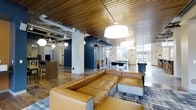 This dynamic amenity space seamlessly combines social and academic environments within a single, thoughtfully designed floor. The club room anchors the space with a bold aesthetic, featuring a striking warm-toned slatted wood ceiling that contrasts beautifully against exposed industrial ductwork and pristine white structural columns. Rich navy blue accent walls add depth and sophistication to the open layout.

At the heart of the lounge area, a generous caramel-toned leather sectional sofa sits atop a patterned area rug, creating an inviting gathering spot beneath oversized globe and drum pendant lights. Beyond the lounge, a pool table positioned near expansive floor-to-ceiling windows takes full advantage of the natural light pouring in and offering views of the surrounding exterior.

Glass-enclosed study rooms line the interior, providing semi-private, distraction-free spaces ideal for focused work or small group collaboration. Each room is visible through transparent walls, maintaining an open and connected feel throughout. A dedicated workstation area is also present, adding further functionality to the space.

The flooring transitions between warm hardwood and polished tile, subtly defining each zone while maintaining a cohesive, modern aesthetic throughout this versatile and well-appointed communal space.