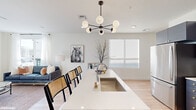 This contemporary single-floor residence showcases a thoughtfully designed open-concept layout that seamlessly connects the kitchen, dining, and living areas. The kitchen is a standout feature, outfitted with bold navy cabinetry, white subway tile backsplash, stainless steel appliances, and a generous white quartz island with an undermount sink and bar seating — all anchored by a striking mid-century modern multi-globe chandelier. Light wood flooring runs throughout, creating a warm, cohesive flow from room to room.

A bright, airy hallway — accented by a gold-framed round mirror — connects the entry to the main living spaces. The living room is bathed in natural light from large windows and a glass-paneled door that opens directly onto a private outdoor balcony, where a wooden dining table and decorative railing frame open neighborhood views under wide skies.

The bedroom offers a serene, minimalist retreat with soft neutral tones, light wood floors, and ample natural light filtering through sheer curtains. The full bathroom features distinctive large hexagonal floor tile, a subway-tiled tub-shower combination, and a dark-toned vanity, balancing classic and modern design elements throughout this well-appointed home.