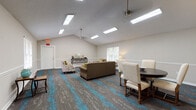 Located at 6655 Mount Zion Blvd in Morrow, Georgia, this single-floor community space offers a welcoming and well-appointed environment designed for comfortable gatherings. The main area is defined by soaring vaulted ceilings fitted with multiple skylights and recessed panel lighting, flooding the room with abundant natural and artificial light. Ceiling fans provide additional comfort throughout the space.

The open layout is thoughtfully arranged to serve multiple functions. A generously sized lounge area features a plush sofa, accent armchairs with colorful throw pillows, and a round wooden coffee table, creating an inviting setting for relaxed conversation. Adjacent to this is a formal dining area with a dark wood round table and elegantly upholstered chairs, suitable for meetings or shared meals. A classic console table along the wall adds a refined decorative touch.

Underfoot, a bold patterned carpet in grey and teal tones adds a vibrant, modern aesthetic that ties the space together. White wainscoting runs along the lower walls, complementing the neutral, light-toned upper walls and contributing to a clean, polished appearance. Large windows offer views of surrounding greenery, enhancing the airy ambiance.

A compact kitchenette off the main area includes white cabinetry, speckled countertops, and a double stainless steel sink with a gold-finish faucet, providing practical support for event catering and everyday use.