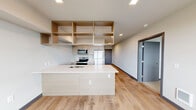 The Aspire 1-bedroom, 1-bathroom residence in Tacoma, WA offers a thoughtfully designed modern living space with a clean, contemporary aesthetic throughout. The open-concept kitchen and living area serve as the heart of the home, highlighted by a large center island with a white countertop and integrated sink, stainless steel appliances including a refrigerator, range, microwave, and dishwasher, and warm wood-toned cabinetry. Open upper shelving adds both storage and a stylish architectural element, while recessed lighting and light wood-plank flooring enhance the bright, airy atmosphere. A window at the end of the living space brings in natural light and outdoor views. The generously sized bedroom features plush carpeting, a large window overlooking the surrounding neighborhood, and ample natural light. Sliding doors lead to a dedicated closet, providing well-organized storage. The bathroom is finished with wood-grain cabinetry, a vanity with a clean countertop, and light hardwood-style flooring. A standout feature is the in-unit stacked washer and dryer, conveniently located adjacent to the bathroom. The unit's neutral color palette, modern fixtures, and efficient layout make it a functional and stylish urban residence.