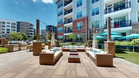 Located at 1055 Hatches Pond Lane in Cary, NC, this well-appointed outdoor courtyard serves as a central amenity space within a modern multi-story residential community. The space is framed by classic brick and contemporary siding facades, creating a visually engaging architectural backdrop that blends traditional and modern design elements.

The courtyard is anchored by a spacious stone-paved terrace that flows throughout the space, connecting several distinct activity zones. A standout feature is the circular outdoor lounge area, centered around a brick fire pit column and furnished with curved wicker seating and vibrant cushions — ideal for social gatherings. Adjacent to this is a well-equipped outdoor kitchen and bar area with brick detailing, providing a functional space for entertaining.

Multiple raised garden beds, constructed in both wood and composite materials, are thoughtfully distributed throughout the courtyard, offering residents the opportunity to cultivate plants and produce. The landscaping throughout is lush and carefully maintained, with ornamental grasses, evergreen shrubs, flowering plants, and mature trees adding seasonal color and texture.

A more intimate seating area tucked near the building features wooden benches surrounded by curated plantings and large-scale container gardens, offering a quiet retreat. The space benefits from abundant natural sunlight and open sky views, creating a bright and welcoming outdoor environment year-round.
