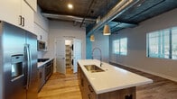 This single-floor unit showcases a striking blend of modern design and industrial aesthetics. The open-concept layout is anchored by a well-appointed kitchen featuring a large center island with a quartz countertop, a deep stainless steel sink with a commercial-style faucet, and two-tone cabinetry combining white upper cabinets with wood-grain lower units. Stainless steel appliances include a French door refrigerator with ice and water dispenser, a gas range, a wall oven, and a built-in microwave, all complemented by a subtle tile backsplash. Exposed silver ductwork runs prominently across the darkened ceiling, adding to the unit's industrial character, while pendant lighting over the island provides focused illumination. The living area extends generously from the kitchen, bathed in natural light through large windows fitted with horizontal blinds. Warm hardwood-style flooring flows consistently throughout the main living areas, transitioning to a richer wood tone in the bedroom corridor. A dedicated laundry closet houses a stacked washer and dryer. The bathroom is cleanly finished with a quartz-topped vanity, wood-grain cabinetry, an LED-backlit mirror, and a full tub-shower combination with white tile surround.