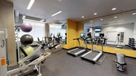 Located at 206 Bell St in Seattle, WA, this fitness center offers a well-equipped, single-floor space designed to support a comprehensive workout routine. The gym features a modern, functional layout with a neutral color palette accented by bold yellow walls that add energy to the environment. Recessed and linear lighting throughout ensures the space is consistently well-lit, while large windows along one wall invite natural light and provide views of the surrounding area.

The cardio zone is outfitted with multiple treadmills and elliptical machines, each equipped with individual digital display screens for interactive workout tracking. Full-length mirrors span a significant portion of the walls, visually expanding the space and allowing users to monitor their form during exercise. The strength training area includes a cable and weight machine, a variety of stability balls, and two organized dumbbell rack towers offering a range of free weights.

Additional amenities include a water cooler station, neatly stored towels, a utility sink, and a mounted flat-screen television for entertainment during workouts. The rubber-textured flooring throughout provides durability and safety underfoot. Climate control is handled via a ceiling-mounted HVAC unit, ensuring year-round comfort. The overall atmosphere is clean, professional, and purpose-built for effective fitness training.