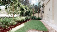 This single-floor space is distinguished by its beautifully maintained outdoor grounds, which showcase a vibrant tropical landscape. The exterior features a well-kept lawn bordered by rich, red-mulched garden beds filled with a variety of tropical plants, including tall palm trees, broad-leafed shrubs, and dense greenery that create a layered, lush aesthetic. The contrast between the manicured grass, the dark mulch, and the light gravel accents along the perimeter gives the outdoor area a polished, intentional design. A cream-toned building facade runs alongside the garden, complementing the natural surroundings with a clean, neutral finish. Mature trees in the background provide ample shade and a sense of privacy, while the bright, open sky above allows generous natural light to filter through the canopy. The overall ambiance of the outdoor space is serene and well-tended, evoking a resort-like atmosphere suited to a warm, subtropical climate. The interior images provided are too dimly lit to yield detailed observations, though the presence of doorways and room transitions suggests a functional interior layout. Together, the grounds and structure present a space that prioritizes pleasant outdoor living within a carefully landscaped, tropical setting.