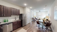Located at 1101 Pure St in Saint Peters, Missouri, this single-floor unit offers a thoughtfully designed open-concept layout that seamlessly connects the kitchen, dining, and living areas. The kitchen is equipped with rich dark espresso cabinetry, granite countertops, a subway tile backsplash, and a full suite of stainless steel appliances, including a refrigerator, range, microwave, and dishwasher. Wood-style plank flooring runs throughout the main living areas, providing a warm and cohesive aesthetic. The dining area accommodates a round table with comfortable seating, positioned conveniently between the kitchen and living room. The living room is bright and inviting, featuring a large window that allows natural light to fill the space, complemented by a ceiling fan overhead. The bedroom continues the warm tone of the unit, with a ceiling fan, a generously sized window with outdoor views, and ample room for a full furniture arrangement. The bathroom features matching dark cabinetry, a granite-topped vanity, a tub-shower combination, and the same wood-style flooring found throughout the unit. Neutral wall tones and recessed lighting create a clean, modern ambiance across all areas of this well-designed home.
