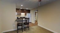 This single-floor space offers a practical and comfortable layout designed for efficient everyday living. The open-concept design seamlessly connects the kitchen and living area, creating a cohesive and functional environment. The kitchen is equipped with rich, dark wood cabinetry that provides ample storage, complemented by under-cabinet lighting that adds warmth to the space. A white refrigerator and a range contribute to a clean, classic look. A built-in breakfast bar with two dark wood bar stools serves as a natural divider between the kitchen and the carpeted living area, making it an ideal spot for casual dining or conversation. Recessed lighting throughout the kitchen ensures the space is well-lit and inviting. The flooring transitions from warm-toned hardwood in the kitchen and hallway to plush carpet in the living area, adding a cozy texture underfoot. Neutral beige walls throughout provide a versatile backdrop that suits a variety of décor styles. A ceiling fan is visible in the living area, offering both comfort and airflow. A hallway visible from the main living space leads to a bathroom, which features a shower with a curtain and standard fixtures, completing this well-organized and functional layout.