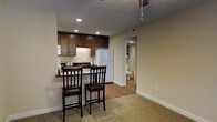 This single-floor space offers a practical and comfortable layout designed for efficient everyday living. The open-concept design seamlessly connects the kitchen and living area, creating a cohesive and functional environment. The kitchen is equipped with rich, dark wood cabinetry that provides ample storage, complemented by under-cabinet lighting that adds warmth to the space. A white refrigerator and a range contribute to a clean, classic look. A built-in breakfast bar with two dark wood bar stools serves as a natural divider between the kitchen and the carpeted living area, making it an ideal spot for casual dining or conversation. Recessed lighting throughout the kitchen ensures the space is well-lit and inviting. The flooring transitions from warm-toned hardwood in the kitchen and hallway to plush carpet in the living area, adding a cozy texture underfoot. Neutral beige walls throughout provide a versatile backdrop that suits a variety of décor styles. A ceiling fan is visible in the living area, offering both comfort and airflow. A hallway visible from the main living space leads to a bathroom, which features a shower with a curtain and standard fixtures, completing this well-organized and functional layout.