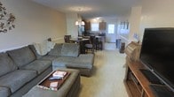 This single-story home in Greenville, North Carolina offers an open-concept floor plan that seamlessly connects the main living areas. The spacious living room is carpeted and anchored by a large sectional sofa and ottoman, creating a comfortable gathering space. A wooden media console adds a traditional touch along one wall.

The living area flows naturally into the dining space, where a round dark-wood table with matching chairs provides an inviting spot for meals. Just beyond, the kitchen is outfitted with medium-toned wood cabinetry, dark countertops, and standard appliances including a refrigerator. A window above the sink allows natural light to filter into the cooking area. An elegant chandelier with candle-style lights hangs above the dining area, adding a decorative focal point overhead.

A secondary entry door with a window is visible at the far end of the kitchen, providing additional access and light. The neutral wall tones and light carpet throughout the main living areas give the home a warm, cohesive feel. The single-floor layout offers convenient, accessible living in a welcoming residential setting.