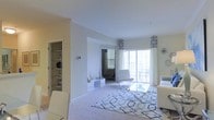 This single-floor residence offers a clean, contemporary design with a bright and airy ambiance throughout. The living area is the centerpiece of the space, showcasing plush carpeted flooring and a large geometric-patterned area rug in navy and white that anchors the room. Modern white furnishings, including sleek seating and a glass-top coffee table, contribute to a polished, minimalist aesthetic. Large sliding glass doors flood the room with natural light and provide access to an outdoor area, framed by elegant patterned window valances in a navy geometric print that add a refined decorative touch. The neutral, warm-toned walls and smooth ceilings enhance the sense of openness throughout. A built-in shelving unit near the entryway offers practical display and storage space, complementing the overall layout. An arched passageway connects the living area to an adjacent room, adding architectural character and maintaining a seamless flow between spaces. The thoughtful combination of natural light, open sightlines, and coordinated design details creates a welcoming and well-appointed environment suited for comfortable everyday living.