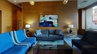 Located at 14 Easton Ave in New Brunswick, NJ, this single-floor lobby space offers a refined and welcoming first impression through a thoughtfully designed interior. The space is defined by its striking horizontal wood-paneled walls, which lend warmth and a contemporary aesthetic throughout. Large floor-to-ceiling windows flood the lounge area with natural light while offering clear views of the surrounding streetscape, creating a seamless connection between the interior and the urban environment outside.

The lounge area is furnished with plush navy blue sofas and accent chairs arranged around a glass-top coffee table, complemented by decorative pendant lighting with gold geometric fixtures and coordinating table lamps. A bold piece of abstract wall art anchors the seating area, adding a vibrant artistic focal point. The tiled floors feature subtle inlaid accents that enhance the overall design.

Transitioning deeper into the lobby, a corridor clad in rich dark tile leads to an elevator bay finished with brushed metal doors and smooth dark wall panels. A decorative console table with a glass top and a sculptural sunburst mirror add refined touches near the main circulation path. The space also includes a fire command center alcove, highlighting its functional, well-equipped infrastructure. Overall, the lobby exudes a polished, modern ambiance suited to a upscale residential or mixed-use building.