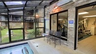 This single-floor amenity space features an enclosed spa and pool area designed for year-round use. The room is defined by expansive floor-to-ceiling glass walls and a glass ceiling structure framed in dark metal, allowing natural light to filter in while providing clear sightlines to the surrounding landscaped courtyard. The exposed brick walls add a warm, textural contrast to the sleek glazing system, lending the space a grounded, welcoming character.

The spa area includes a built-in hot tub or plunge pool with tiled surround and step-in access, finished with neutral, durable poolside flooring throughout. A pair of wrought iron chairs provides seating adjacent to the water, suitable for relaxing before or after use. Warm overhead lighting complements the natural light, ensuring the space remains well-illuminated at any hour.

A sliding glass door connects this area to an adjacent interior room visible through the window, which appears to contain fitness equipment. Safety signage, including spa rules and emergency contact information, is posted throughout, reflecting the space's commitment to user safety. Overall, the area offers a functional and serene recreational environment.