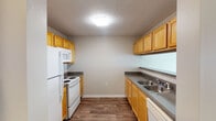 Located at 601 Friendway Rd in Greensboro, NC, this single-floor residential unit offers a practical and well-organized layout designed for comfortable everyday living. The galley-style kitchen is efficiently arranged with warm honey-toned wood cabinetry lining both sides, dark countertops, wood-look flooring, and white appliances including a refrigerator, electric range, built-in microwave, and dishwasher. A double basin sink with ample counter space adds to the kitchen's functionality. The open living area features light hardwood-style flooring and large windows that invite natural light, creating a bright and airy atmosphere. A pendant light fixture with a classic chandelier design adds a decorative touch near the kitchen pass-through. The bedroom is finished with plush carpeting, a ceiling fan with light, and a window with blinds providing natural light and ventilation. Doors from the bedroom lead to a bathroom and additional spaces, offering a connected and private feel. The bathroom includes a full soaking tub and shower combination with a curtain rod, wood-look flooring, and clean neutral finishes throughout. Textured ceilings are present throughout the unit, and neutral wall tones lend a calm, cohesive ambiance to the entire space.