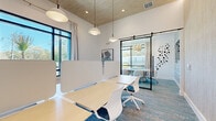 Located at 8707 N Davis Hwy in Pensacola, FL, this contemporary single-floor coworking space offers a thoughtfully designed, productivity-focused environment. The open workspace features light wood benching workstations fitted with white privacy screens, complemented by modern pendant dome lighting that hangs from a striking wood-paneled ceiling. Large, black-framed floor-to-ceiling windows flood the space with natural light and offer views of the lush, tree-lined surroundings outside.

A glass-walled conference room, accessible through sleek black barn-style sliding doors, provides a semi-private meeting area equipped with a wooden conference table, upholstered chairs, and a wall-mounted display screen. The conference room's accent wall features a textured, horizontal wood-look finish, adding warmth and visual interest. An abstract sculptural wall installation adds an artistic touch near the glass partition.

The space is completed by multi-toned blue and grey patterned carpeting throughout, white walls, and recessed lighting that enhances the bright, airy ambiance. The overall design strikes a balance between open collaboration and focused privacy, making it a versatile and professional workspace.