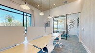Located at 8707 N Davis Hwy in Pensacola, FL, this contemporary single-floor coworking space offers a thoughtfully designed, productivity-focused environment. The open workspace features light wood benching workstations fitted with white privacy screens, complemented by modern pendant dome lighting that hangs from a striking wood-paneled ceiling. Large, black-framed floor-to-ceiling windows flood the space with natural light and offer views of the lush, tree-lined surroundings outside.

A glass-walled conference room, accessible through sleek black barn-style sliding doors, provides a semi-private meeting area equipped with a wooden conference table, upholstered chairs, and a wall-mounted display screen. The conference room's accent wall features a textured, horizontal wood-look finish, adding warmth and visual interest. An abstract sculptural wall installation adds an artistic touch near the glass partition.

The space is completed by multi-toned blue and grey patterned carpeting throughout, white walls, and recessed lighting that enhances the bright, airy ambiance. The overall design strikes a balance between open collaboration and focused privacy, making it a versatile and professional workspace.