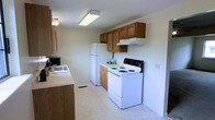 This two-floor residential space offers a straightforward and functional layout designed to meet everyday living needs. The kitchen is a key feature of the home, showcasing warm honey-toned wood cabinetry that provides ample storage both above and below the counter. The space is equipped with essential appliances, including a white refrigerator, an electric coil range with an overhead ventilation hood, and a sink integrated into the countertop. Light-toned counters and vinyl flooring contribute to a clean, easy-to-maintain aesthetic throughout the kitchen. Overhead fluorescent lighting ensures the workspace is well-lit, making meal preparation convenient. The neutral wall tones create a bright and open atmosphere, allowing the cabinetry to serve as the room's defining visual element. A floor vent indicates a central heating system, adding to the practicality of the space. Spanning two floors, this home provides separation between living areas, offering both communal and private spaces across its levels. The overall ambiance is modest and approachable, with an emphasis on functionality and comfortable, day-to-day usability.