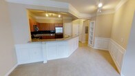 This single-floor residence showcases an open-concept layout that seamlessly connects the kitchen and living area, creating a spacious and cohesive environment. The kitchen is outfitted with warm-toned wood cabinetry, dark granite countertops, and a classic black tile backsplash that adds contrast and texture. Stainless steel appliances, including a built-in double oven and refrigerator, contribute a modern, polished look. A generously sized kitchen island with a white paneled base and granite surface serves as a natural dividing point between the kitchen and the adjoining living space, offering both functionality and visual appeal.

Crown molding runs throughout the space, adding an elegant architectural detail that elevates the overall design. Recessed track lighting illuminates the kitchen area, while the living space benefits from soft, neutral-toned carpeting that creates a comfortable atmosphere. A set of built-in shelving units with white trim is integrated into the living area wall, providing both display and storage options. Additional doors along the hallway suggest further rooms beyond the main living area. The warm beige tones used throughout lend the space a cohesive and inviting ambiance.