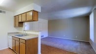 This single-floor space presents an open-concept layout where the kitchen and living area connect naturally, creating a functional and straightforward floor plan. The kitchen is equipped with warm wood-toned cabinetry, both upper and lower units, providing ample storage space. A countertop with an integrated sink anchors the kitchen area, which features hard-surface flooring that transitions smoothly into the carpeted living area beyond.

A half-wall partition subtly defines the boundary between the kitchen and the living space without fully closing off the two areas, maintaining an open and connected feel. The living area is finished with wall-to-wall carpet and neutral-toned walls, offering a clean, blank canvas. A floor vent is visible along the baseboard, indicating a built-in climate control system.

The textured ceiling runs throughout the space, and the overall color palette is understated and neutral, emphasizing practicality. A closet with bifold-style doors is visible in the background, adding dedicated storage. The layout is efficient and straightforward, suited to everyday living.