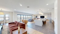 Located at 2 Rivers Run Dr in Schenectady, NY, this thoughtfully designed single-floor community space offers a generous, open-concept layout that seamlessly blends multiple functional areas. The main gathering area is anchored by a spacious living zone featuring rich leather seating, a large-format wall-mounted television set against a striking marble-patterned accent wall, and warm wood flooring that flows throughout. Recessed lighting and globe pendant fixtures provide layered illumination, enhancing the clean, contemporary aesthetic defined by bright white walls and smooth ceilings.

A central bar-height island with seating serves as a natural divider between the lounge and dining areas, while an expansive wall of floor-to-ceiling dark-framed windows and sliding glass doors floods the interior with natural light and provides direct access to an outdoor balcony with sweeping views of the surrounding landscape. A dedicated game room offers an intimate retreat, centered around an octagonal game table encircled by plush upholstered chairs, bathed in natural light from a trio of black-framed windows and accented by decorative wallcovering. Together, these spaces create a versatile, well-appointed environment ideal for social gatherings and everyday leisure.