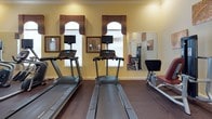 This fitness center offers a comprehensive workout environment designed to accommodate a variety of exercise routines. The space is finished with warm yellow walls, white crown molding, and a rich tray ceiling accented by a ceiling fan, lending the room an inviting, polished aesthetic that sets it apart from a typical gym setting.

The cardio section features two treadmills, each equipped with individual flat-screen monitors, as well as an elliptical trainer, providing ample options for aerobic exercise. A dedicated strength training area includes multiple cable and resistance machines, a flat utility bench, and a well-stocked dumbbell rack offering a range of free weights. Foam rollers and additional accessories are also available for stretching and recovery.

Large wall-mounted mirrors throughout the space enhance the sense of openness and allow users to monitor their form during workouts. Natural light enters through several arched windows dressed with decorative valances, contributing to the bright and energizing atmosphere. A wall-mounted television and clock are centrally positioned for easy viewing. Durable rubber flooring covers the entire area, providing both safety and comfort. Access is provided through a glass-paned door, connecting the fitness center to adjacent amenity spaces.