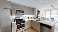 This thoughtfully designed single-floor space showcases a clean, contemporary aesthetic that is both functional and visually appealing. The kitchen serves as a standout feature, outfitted with a full suite of stainless steel appliances, including a gas range, over-the-range microwave, and dishwasher. Flat-front cabinetry in a warm taupe wood-grain finish extends floor to ceiling, maximizing storage while contributing to the space's modern character. Crisp white quartz countertops provide ample prep space and contrast beautifully against the textured mosaic tile backsplash, which adds depth and visual interest to the cooking area. A stainless steel undermount sink is positioned at the peninsula, which opens toward the living area, creating a natural flow between spaces and making the layout ideal for both everyday living and entertaining. Warm-toned hardwood flooring grounds the space with a natural element, complementing the neutral color palette found throughout. Recessed lighting and an abundance of natural light contribute to a bright, airy ambiance. A lush indoor plant near the living area adds a refreshing touch of greenery, enhancing the overall sense of warmth and livability within this well-appointed space.