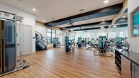 This well-appointed fitness center offers a generously sized, single-floor layout designed to accommodate a full range of workout activities. The space is finished with warm-toned hardwood-style flooring throughout, creating a clean and inviting atmosphere that complements the bright, modern interior.

Architecturally, the facility stands out with bold dark-painted exposed ceiling beams that add a contemporary industrial character, while recessed lighting and ceiling fans ensure consistent illumination and comfortable airflow. Natural light pours in through large windows and glass-panel entry doors, providing views of the surrounding outdoors and contributing to an energizing environment.

The equipment selection is extensive and thoughtfully arranged. Cardio offerings include treadmills, stair climbers, stationary bikes, and elliptical machines, all distributed to allow ample space between users. A dedicated strength training zone houses a variety of cable machines and resistance equipment, while a separate functional training area features a well-stocked rack with kettlebells, dumbbells, stability balls, resistance tools, and a plyo box—ideal for dynamic and free-weight workouts.

Accent walls in warm terracotta tones and natural wood paneling add visual warmth, while large mirrors enhance the sense of openness. The overall design balances functionality with aesthetics, creating a motivating and professional fitness environment.