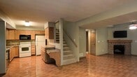 This two-story residential space offers a practical and welcoming layout across both levels. The main floor showcases an open-concept design where the kitchen flows naturally into the living area, creating a connected and versatile living environment. The kitchen is equipped with warm-toned wood cabinetry, a white refrigerator, a built-in microwave, and a peninsula countertop that provides additional workspace and casual seating potential. The countertops feature a stone-look finish that complements the overall color palette. Light wood-style flooring runs throughout the main level, adding warmth and continuity to the space. The walls are finished in a soft, neutral gray tone, contributing to a calm and understated ambiance. A textured ceiling adds a classic residential character throughout. A carpeted staircase with a wooden handrail leads to the upper floor, providing a smooth transition between levels. The layout also includes a doorway leading to additional rooms or utility spaces, enhancing the overall functionality of the home. The space is well-suited for everyday living, with a straightforward floor plan that balances openness with defined areas for cooking, dining, and relaxing.