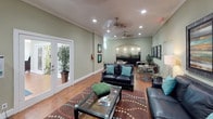 This inviting clubroom is designed to offer a versatile and comfortable gathering space. The main lounge area is anchored by a dark leather sofa and loveseat accented with teal throw pillows, arranged around a glass-top coffee table atop a richly patterned area rug. Warm hardwood flooring extends throughout, complementing the soft sage green walls adorned with colorful framed artwork. Recessed lighting and ceiling fans with sleek metallic finishes provide both illumination and comfort across the space. A large entertainment center with a flat-screen television serves as a focal point in the lounge, complete with shelving for décor and media. Toward the back of the room, a kitchenette or wet bar area is highlighted by a tasteful tile backsplash and under-cabinet lighting, accompanied by high-top seating for casual dining or socializing. A separate bistro-style table with chairs offers additional seating. French doors along one wall flood the interior with natural light and provide access to an adjacent outdoor area. Crown molding and vaulted ceiling details add an architectural refinement that elevates the overall ambiance of this functional and stylish community space.