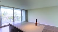 This single-floor space showcases a clean, contemporary design with an open-concept layout that seamlessly connects the kitchen and living areas. The kitchen features a generous white quartz countertop island with a smooth, polished surface, complemented by dark cabinetry underneath that provides a striking contrast. The minimalist aesthetic is reinforced by the bright, neutral wall tones and light-toned flooring that runs throughout the space.

A standout feature is the expansive floor-to-ceiling sliding glass doors that flood the interior with natural light and open directly onto a private balcony. The balcony offers outdoor views of surrounding greenery and neighbouring architecture, creating a pleasant connection between the interior and exterior. Vertical blinds flanking the sliding doors allow for adjustable light control and privacy.

The living area is open and versatile, providing ample space for furniture arrangement. The smooth, light-toned ceiling adds to the airy feel of the space. Overall, the unit presents a bright, modern ambiance with thoughtful design details that balance functionality and style, making it a comfortable and visually appealing living environment.