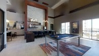 This single-floor amenity lounge is designed to impress, combining contemporary aesthetics with functional social spaces. The room's most striking feature is its soaring ceiling height, which amplifies the sense of openness and grandeur throughout. A dramatic floor-to-ceiling textured stone accent wall, framed by rich wood trim, anchors the space and serves as a stunning focal point.

The lounge centers around a sleek billiards table positioned atop a boldly patterned area rug in earth tones of rust, navy, and gray, adding a dynamic visual element to the light-toned flooring. Adjacent to this, a bar or lounge counter area is outfitted with dark seating and floating shelves that display decorative accents, illuminated by a cluster of pendant lights that add warm, ambient lighting.

Natural light filters in through a large window, contributing to the bright and welcoming atmosphere. A mirror on the opposing wall reflects light and enhances the sense of depth. The overall ambiance balances upscale, modern design with a relaxed, social environment suited for gathering and recreation.