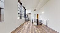 This two-story space combines industrial character with modern finishes throughout. The upper level serves as the main living area, immediately distinguished by soaring ceilings and dramatic floor-to-ceiling industrial-style windows with bold black frames. These oversized multi-pane windows flood the space with abundant natural light and offer views of the surrounding streetscape. Warm wood-look plank flooring runs continuously throughout both levels, lending a cohesive, contemporary feel. A sleek black metal railing frames the open staircase, connecting the two floors with an architectural flair.

The kitchen is efficiently laid out in a galley configuration, featuring gray cabinetry, granite-look countertops, a subway tile backsplash, and stainless steel appliances including a refrigerator and range. A bedroom on the lower level is generously proportioned and features a closet and access to a clean, well-lit bathroom complete with a soaking tub, shower, vanity, and wood-look flooring. An additional half bath with a white vanity and mirror provides added convenience. Throughout the space, crisp white walls and neutral tones create a bright, open ambiance.