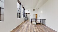 This two-story space combines industrial character with modern finishes throughout. The upper level serves as the main living area, immediately distinguished by soaring ceilings and dramatic floor-to-ceiling industrial-style windows with bold black frames. These oversized multi-pane windows flood the space with abundant natural light and offer views of the surrounding streetscape. Warm wood-look plank flooring runs continuously throughout both levels, lending a cohesive, contemporary feel. A sleek black metal railing frames the open staircase, connecting the two floors with an architectural flair.

The kitchen is efficiently laid out in a galley configuration, featuring gray cabinetry, granite-look countertops, a subway tile backsplash, and stainless steel appliances including a refrigerator and range. A bedroom on the lower level is generously proportioned and features a closet and access to a clean, well-lit bathroom complete with a soaking tub, shower, vanity, and wood-look flooring. An additional half bath with a white vanity and mirror provides added convenience. Throughout the space, crisp white walls and neutral tones create a bright, open ambiance.