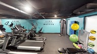 This fitness center in Dallas offers a comprehensive workout environment designed to accommodate a wide range of exercise routines. The space is defined by its bold teal accent walls, which are punctuated by the motivational phrase "Mind Over Matter," creating an energizing atmosphere for members. The exposed ceiling with visible ductwork and dark overhead finishes lend the space a distinctly industrial character, complemented by rubber flooring throughout that ensures durability and comfort during workouts.

The cardio section is well-stocked with treadmills, elliptical machines, and a stair climber, all arranged to allow ample room for movement. A strength training area features a cable machine, an adjustable weight bench, and a full dumbbell rack, catering to both beginner and advanced lifters. Functional fitness accessories are also well-represented, including kettlebells mounted on a wall rack, medicine balls, stability balls, and resistance bands, supporting dynamic and varied training styles.

A mounted flat-screen television, a water cooler station, and mirrored walls add practical touches that enhance the overall experience. The space is organized efficiently, making it easy to transition between different types of training. The cohesive design and thorough equipment selection make this fitness center a highly functional and motivating environment for achieving health and wellness goals.