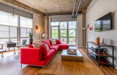 This single-floor loft showcases a striking industrial aesthetic defined by its exposed concrete ceilings, visible steel ductwork, and structural concrete columns throughout. The open-plan living and kitchen area is anchored by generous floor-to-ceiling gridded windows and glass doors that open to a private outdoor balcony, bathing the space in natural light and offering outdoor views. Warm hardwood flooring runs throughout the main living areas, complementing the raw architectural elements above.

The kitchen is well-appointed with granite countertops, a large island with pendant lighting, two-tone cabinetry combining white uppers with dark lower cabinets, a mosaic tile backsplash, and a full suite of stainless steel appliances. The living area benefits from an abundance of natural light sourced from multiple window walls.

The bedroom continues the industrial design language, with exposed pipes and concrete overhead contrasting with soft carpeted flooring and large windows fitted with Roman shades for light control. A rustic reclaimed wood dresser adds warmth to the space.

The bathroom features tile flooring, a soaking tub with subway tile surround, a granite-topped dark vanity, a large mirror, and a tall storage cabinet, providing a clean and functional finish to the space.