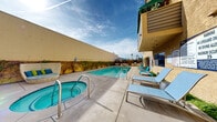 This outdoor recreational area is designed to provide a comprehensive resort-style experience. At the heart of the space is a generously sized rectangular swimming pool with clear, blue-tinted water, complete with lane markings and stainless steel entry rails. Adjacent to the pool sits a circular hot tub, also finished with polished steel handrails, offering a relaxing hydrotherapy option.

The surrounding pool deck is finished in smooth concrete, providing ample space for lounging. A collection of modern light-blue chaise loungers lines the poolside, complemented by additional seating clusters featuring contemporary rattan and woven furniture pieces in teal and navy tones. Blue patio umbrellas are stationed throughout the deck, offering shaded retreats from the sun.

A dedicated outdoor BBQ station is positioned at one end of the deck, equipped with multiple built-in grills and counter space, making it well-suited for outdoor gatherings. Lush ivy climbing along the perimeter walls adds a natural, verdant backdrop to the space.

The area is enclosed by the building's warm yellow stucco exterior, which rises several stories and features balconies overlooking the pool deck. Palm trees visible beyond the enclosure hint at a warm, Southern California-style setting. The overall ambiance is bright, open, and inviting, ideal for both relaxation and social activities.