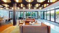 Located in Miami's vibrant Wynwood neighborhood, this expansive amenity and coworking space seamlessly blends contemporary design with lush tropical landscaping. The interior is defined by soaring ceilings with exposed ductwork, polished concrete floors, and floor-to-ceiling glass walls that flood the space with natural light and connect the indoors to the surrounding greenery. Distinctive artistic murals sweep across walls and ceilings, adding bold color and movement to the open-plan layout. A welcoming reception area anchors the main interior zone, complemented by a lounge with a pool table, comfortable seating arrangements, and pendant chandelier lighting. Dedicated coworking areas feature long communal tables, glass-partitioned private offices, and stylishly lit collaboration zones with dark wood ceilings and globe pendants. Multiple outdoor courtyards extend the living and working environment, each paved with mosaic terrazzo tile and surrounded by lush plantings of palms, banana trees, and tropical foliage. A central fire pit lounge with curved white planters and woven outdoor furniture offers a relaxed gathering spot. Walkways with sleek black railings connect the various outdoor zones, creating a cohesive campus-like atmosphere that reflects the energy and creativity of its Miami surroundings.