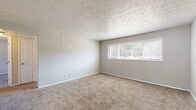 This single-floor unit offers a straightforward, functional layout with multiple rooms spread across a well-organized floor plan. The living areas are finished with neutral gray walls and textured ceilings throughout, creating a consistent, clean aesthetic. Large horizontal windows allow natural light to fill the carpeted rooms, which feature soft, beige wall-to-wall carpeting underfoot.

The common areas and secondary room transition to durable wood-look vinyl flooring in a cool gray tone, offering a practical yet attractive surface. A sliding glass door in one of these rooms provides direct outdoor views and additional natural light, covered by vertical blinds for privacy.

The kitchen is equipped with white cabinetry, light-toned countertops with a subtle speckle finish, a double-basin stainless steel sink, a subway tile backsplash, and a black dishwasher — offering ample storage and workspace.

The bathroom features wood-look tile flooring, a white vanity with under-sink storage, a full tub and shower combination with white tile surround, and a window fitted with horizontal blinds. The overall ambiance of the space is clean, neutral, and move-in ready.