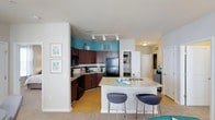 Located at 5721 Goodstone Dr in Raleigh, North Carolina, this single-floor residence offers a thoughtfully designed open-concept layout that seamlessly connects the kitchen and living areas. The kitchen is a standout feature, showcasing rich dark wood cabinetry that extends to the upper walls, complemented by a teal accent wall that adds a vibrant pop of color. Granite countertops provide ample workspace and extend to a curved kitchen island, which doubles as a breakfast bar with seating for two — ideal for casual dining or entertaining. Black appliances, including a refrigerator, dishwasher, and built-in microwave, offer a sleek, cohesive aesthetic. Track lighting overhead ensures the kitchen is well-lit and functional. The open floor plan allows the kitchen to flow naturally into the living space, which features light-toned flooring that enhances the sense of openness. A built-in shelving unit provides convenient storage and display space. The neutral color palette throughout the main living areas, accented by carefully chosen décor, creates a clean and welcoming ambiance. Multiple doorways connect to additional rooms, hinting at a well-organized and practical floor plan designed for comfortable everyday living.