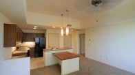 This single-floor residence offers an open-concept layout that seamlessly connects the kitchen and living areas, creating a spacious and functional environment. The kitchen is well-appointed with dark wood cabinetry, a black refrigerator with bottom freezer, and granite countertops that extend across a peninsula with an integrated sink. A secondary island with a warm-toned wood surface provides additional workspace or casual dining options. Two elegant elongated pendant lights hang above the peninsula, adding a decorative focal point while providing focused task lighting. Recessed lighting throughout the ceiling supplements the overall illumination, ensuring the space remains bright and inviting. The kitchen transitions naturally into the carpeted living area, which features a ceiling fan for added comfort. Neutral, warm-toned walls are consistent throughout, contributing to a cohesive and welcoming ambiance. Multiple doorways visible from the main space suggest well-distributed access to additional rooms, enhancing the home's practical flow. The combination of tile flooring in the kitchen and plush carpet in the living area thoughtfully delineates functional zones while maintaining a unified aesthetic throughout.