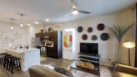 This well-designed two-story residence offers a thoughtfully arranged open-concept living space that seamlessly blends the kitchen and living areas into a cohesive, functional environment. The kitchen is outfitted with stainless steel appliances, including a refrigerator, microwave, and range, all complemented by dark cabinetry with ample storage that extends to the upper cabinets. A light-toned countertop, likely marble or quartz, adds a clean, modern contrast to the rich cabinetry tones. The living area is bright and inviting, featuring wood-look flooring that adds warmth and continuity throughout the space. A ceiling fan with integrated lighting provides both comfort and illumination, while recessed lighting further enhances the airy feel of the room. Decorative woven wall art adds a distinctive, culturally inspired touch to the living space, creating a vibrant focal point alongside the media area. The layout promotes an easy flow between functional zones, making the space ideal for both everyday living and entertaining. With two floors, the residence offers additional space and separation, providing flexibility in how the home is utilized. The overall aesthetic balances contemporary design elements with warm, eclectic accents for a welcoming ambiance.