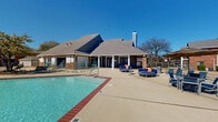 Located at 1400 Sierra Springs Drive in Bedford, Texas, this outdoor amenity complex serves a multi-building residential community spanning three floors. The centerpiece of the space is a generously sized, freeform resort-style swimming pool with crystal-clear blue water, accented by brick coping and complemented by an adjacent spa/hot tub featuring stone surround details. The expansive pool deck is outfitted with a large collection of lounge chairs, ideal for sunbathing, as well as casual seating arrangements including circular conversation sets with cushioned chairs and side tables. A covered cabana area with a pergola-style structure provides shaded outdoor dining and gathering space, furnished with tables, chairs, and a striped market umbrella. The adjacent clubhouse building features a pitched roof with a prominent chimney, brick and siding exterior, and glass-paneled doors opening to the pool area. The community grounds are dotted with mature trees that frame the open-sky views, and winding concrete pathways connect the pool area to the surrounding multi-story brick residential buildings, each featuring private balconies. The overall ambiance is open and resort-like, designed to maximize outdoor leisure and community enjoyment under the expansive Texas sky.